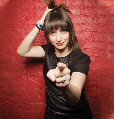 Comedian Ophira Eisenberg performing stand-up in New York City, pointing playfully at the camera against a red brick wall.