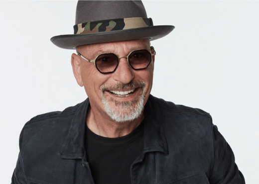Howie Mandel smiling in a stylish hat and sunglasses, wearing a dark jacket