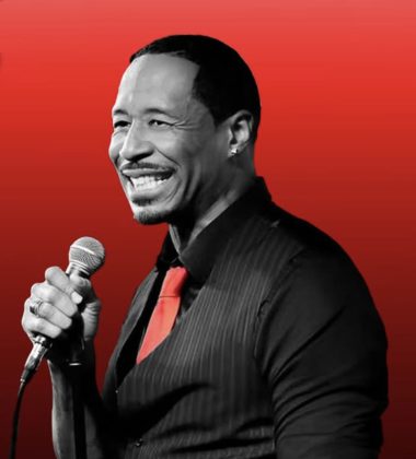 Simeon Goodson performing live at New York Comedy Club in NYC, holding a microphone and smiling on stage.