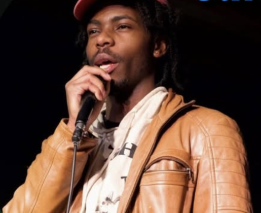 Calder Stokes performing stand-up comedy in New York City, wearing a brown leather jacket and speaking into a microphone on stage.