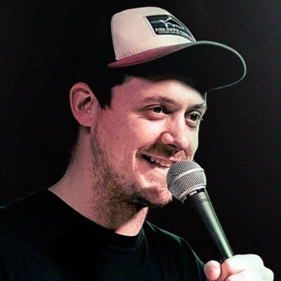 Comedian Colum Tyrrell performing stand-up in New York City, smiling while holding a microphone on stage.